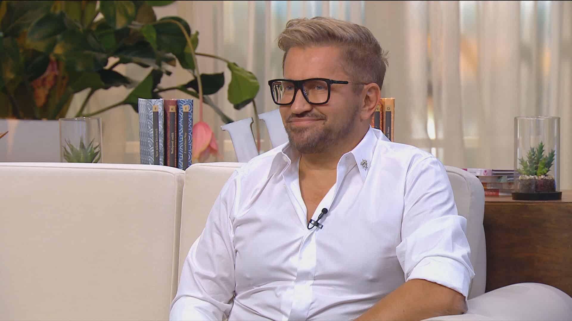 egy férfi fehér ingben és szemüvegben ül egy kanapén egy stúdióban és mosolyog, ez a férfi Jákob Zoltán magyar milliárdos vállalkozó a Crystal Nails tulajdonosa
