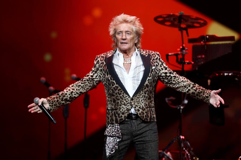 Rod Stewart skót énekes a színpadon lepőrd mintájú felsőben és fehér ingben kócos hajjal