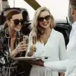 Két mosolygó gazdag nő élvezi a pezsgőt helikopter mellett elegáns férfi társaságában a naplementében
