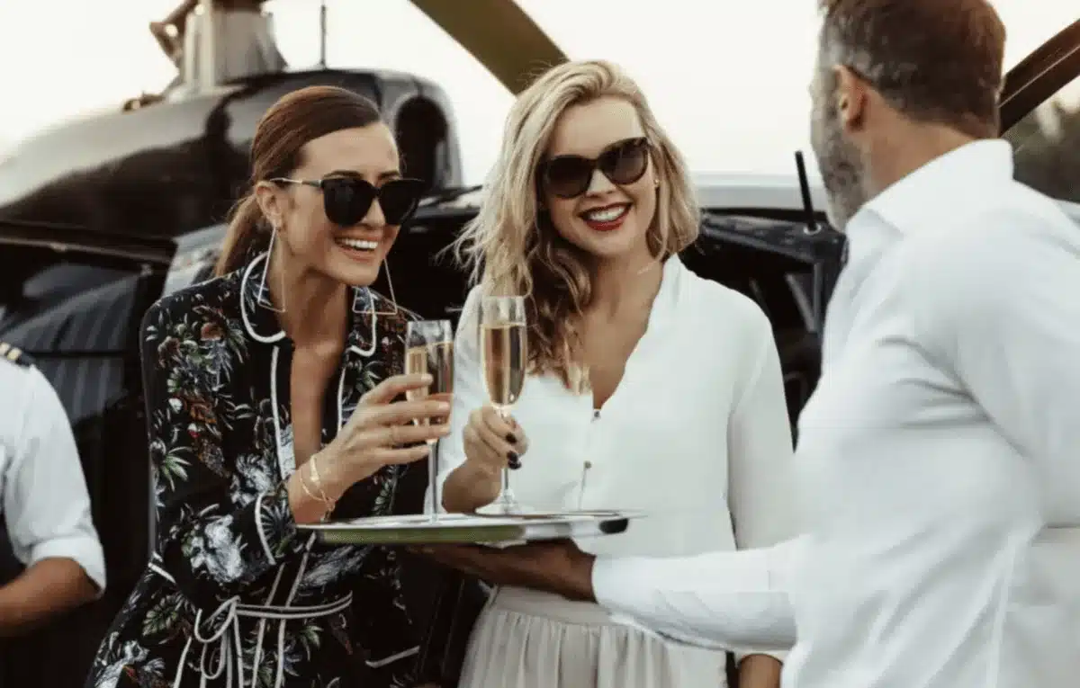Két mosolygó gazdag nő élvezi a pezsgőt helikopter mellett elegáns férfi társaságában a naplementében